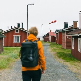 A person wearing an orange jacket and a black backpack walks along a gravel lane between red wooden cottages in Hailuoto, with a windsock visible in the distance.