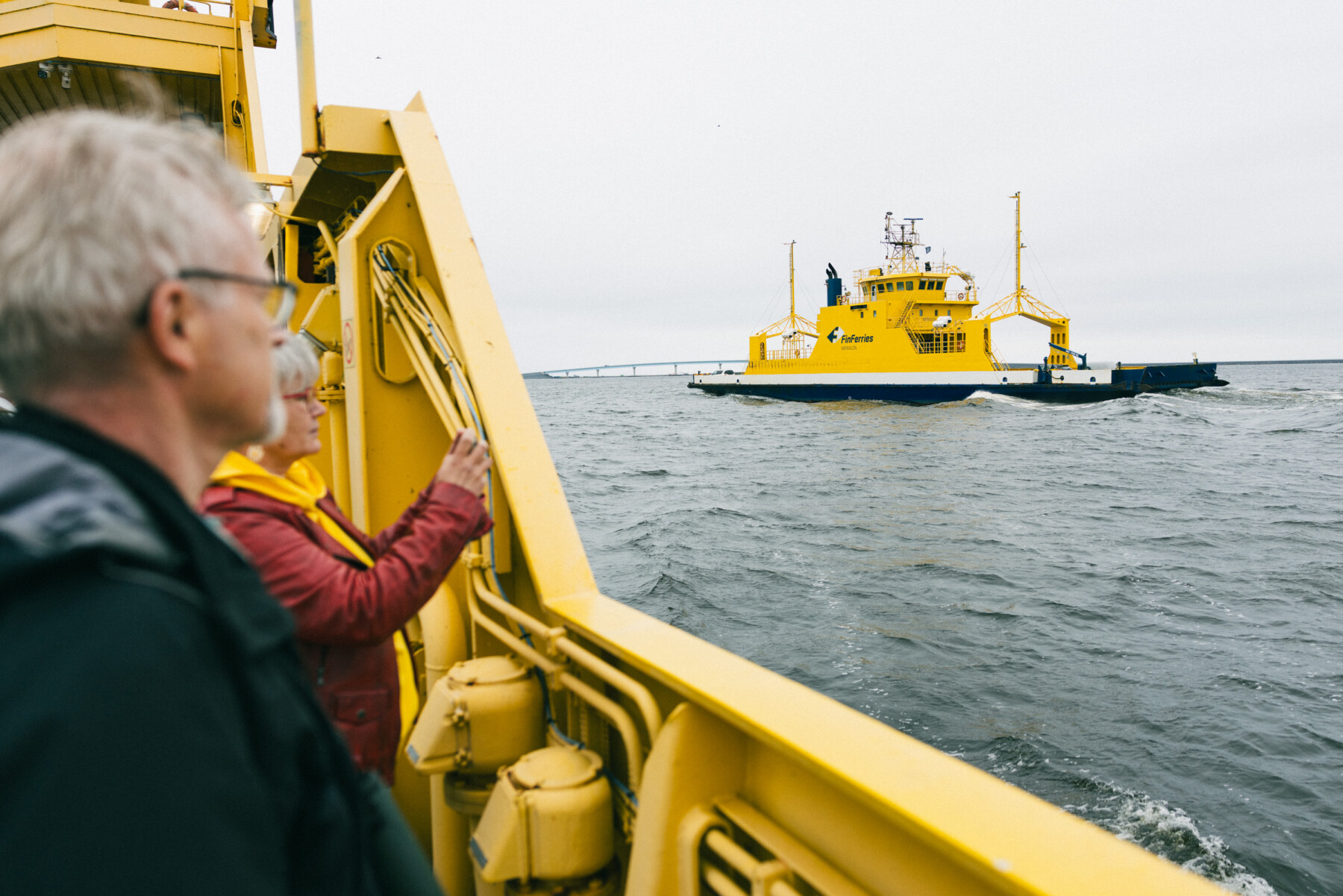Passengers aboard a yellow ferry look out across choppy waters towards a second ferry, with part of the new Hailuoto bridge visible on the horizon.