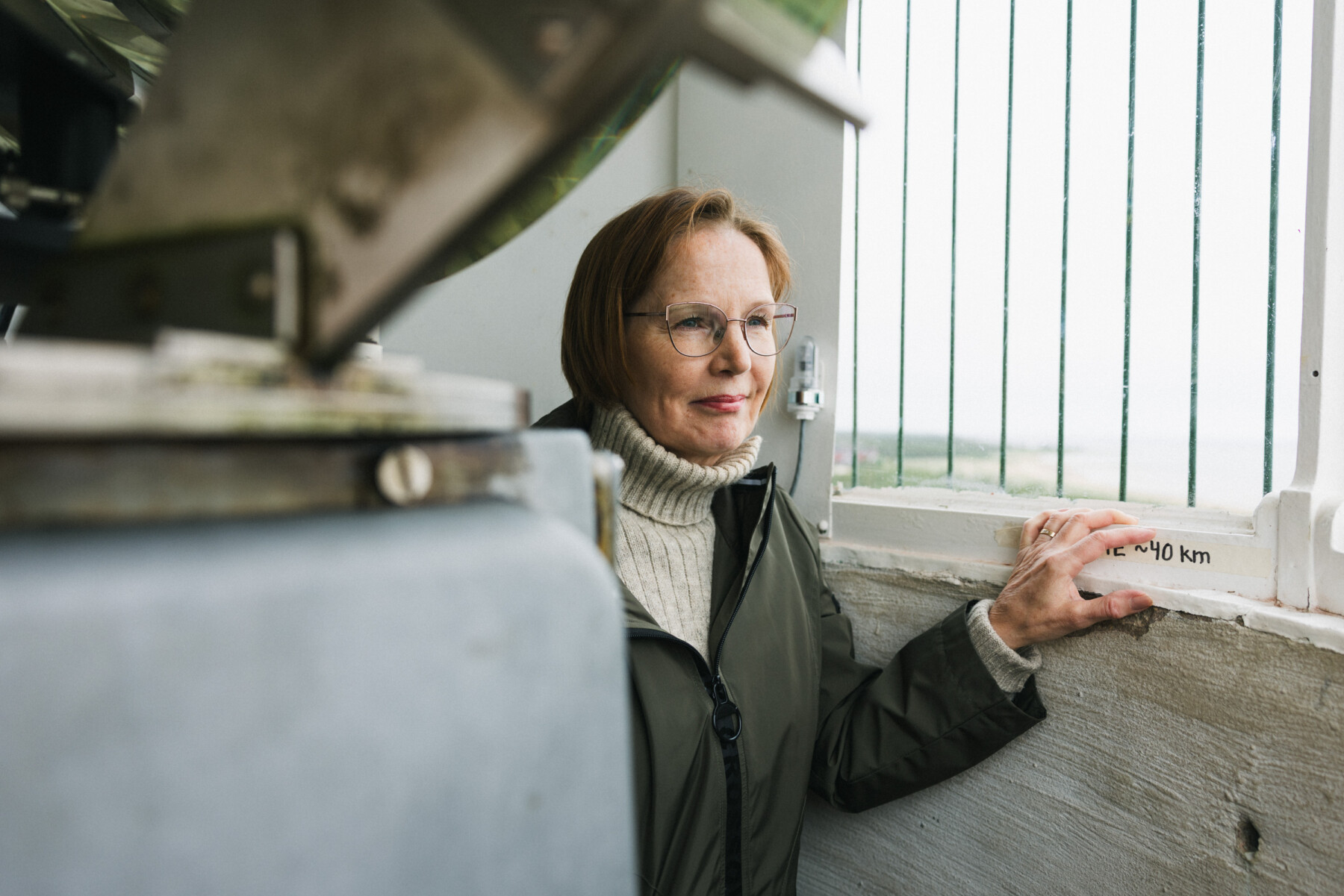 A woman inside Marjaniemi Lighthouse, touches the lantern room window frame beside a distance marker, with the lighthouse lens structure in the foreground.