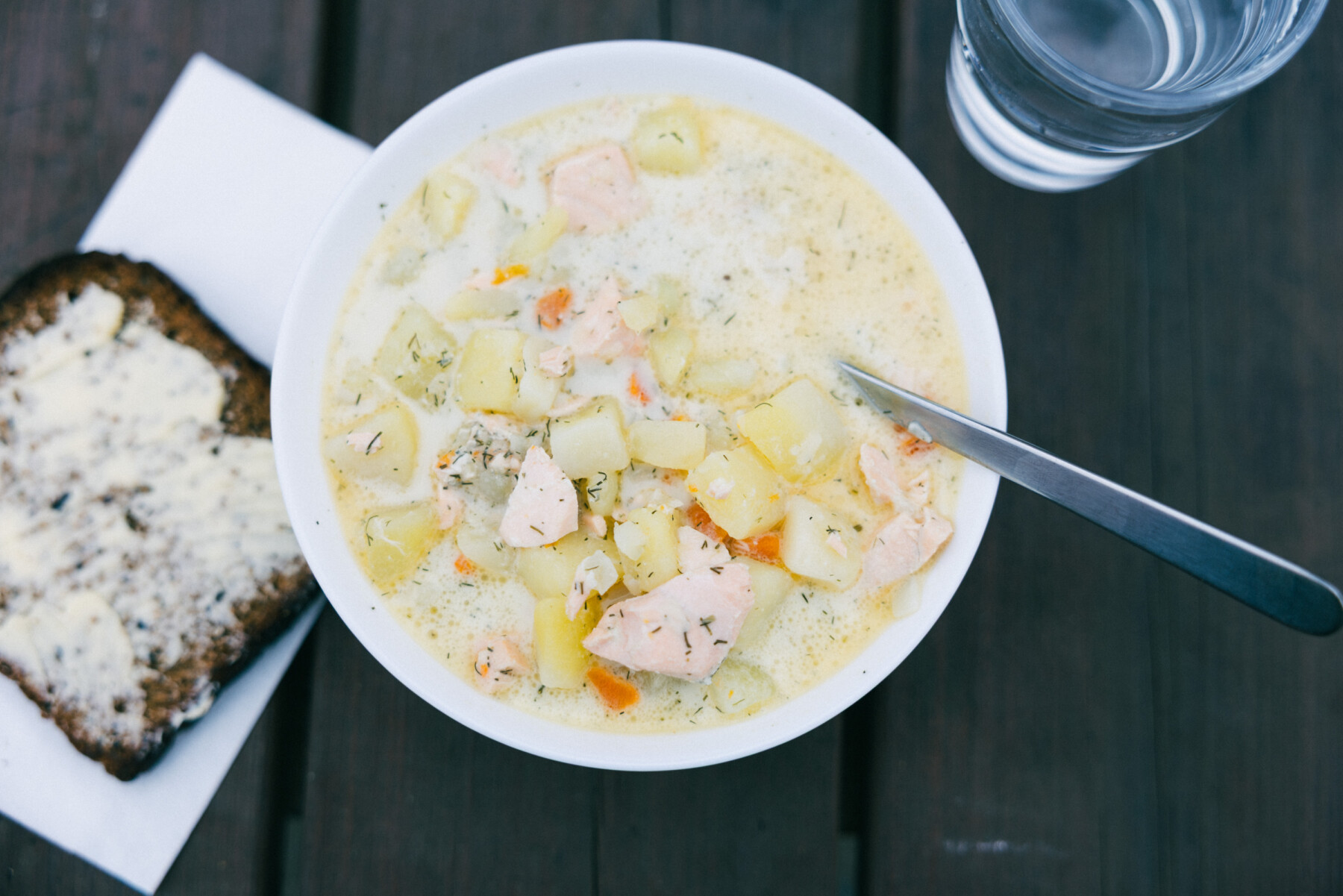 A bowl of creamy salmon soup with diced potatoes and vegetables sits on a dark table beside a slice of buttered rye bread and a glass of water.