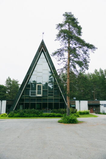 The modernist Hailuoto Church stands amid trees with its steep triangular roof and glass façade visible from the exterior.
