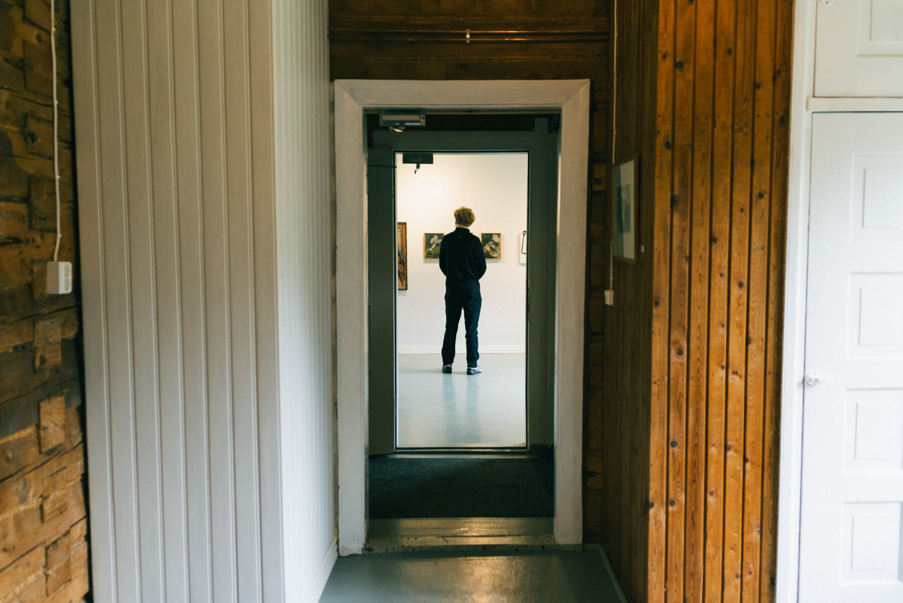 Framed by wooden walls and a narrow corridor, a visitor stands in a brightly lit gallery space, looking at artworks on display at Galleria Luoto in Hailuoto.