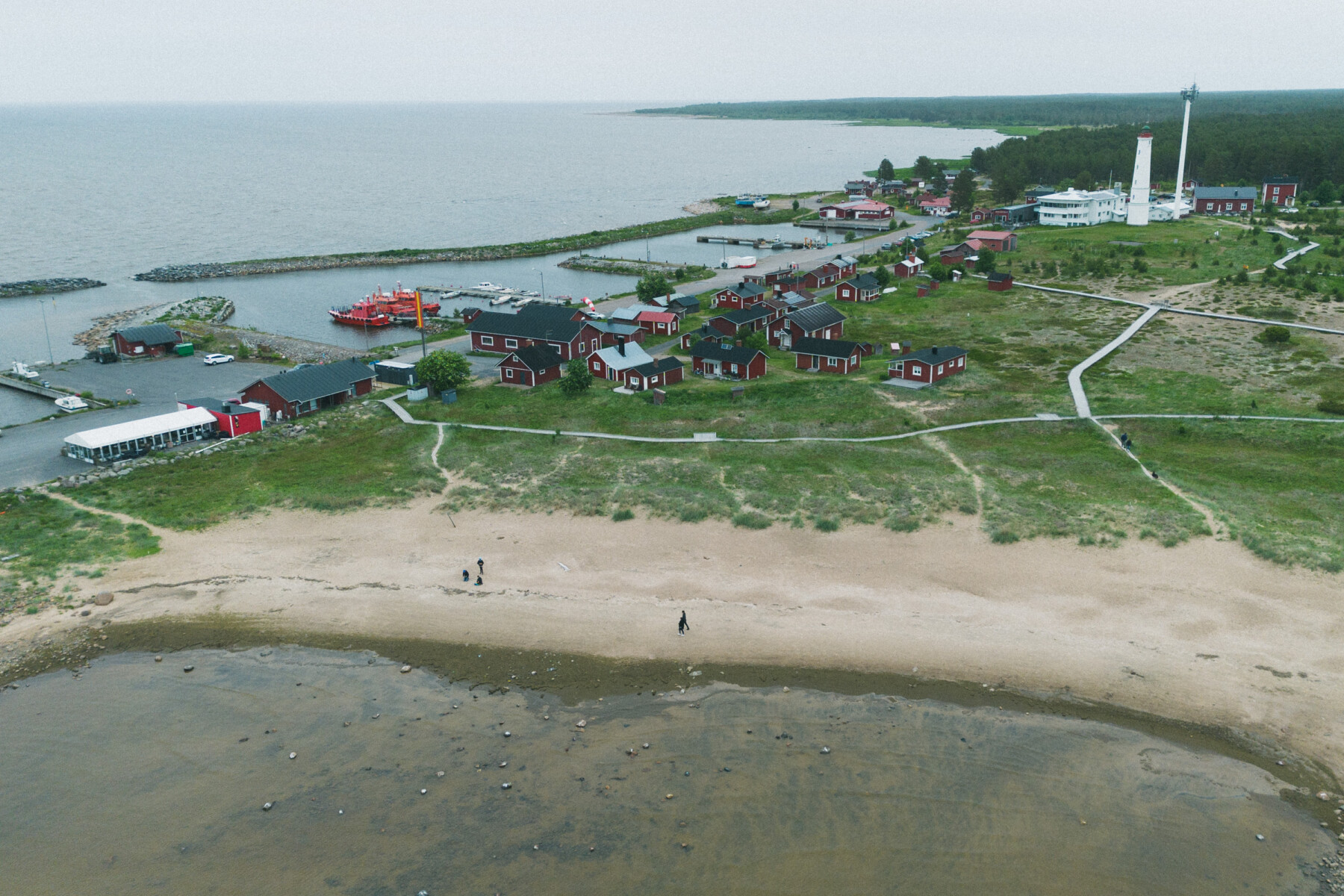 An aerial view shows the shoreline at Marjaniemi on Hailuoto, with sandy beaches, red wooden cottages, a harbour and the Marjaniemi Lighthouse beside the Gulf of Bothnia.