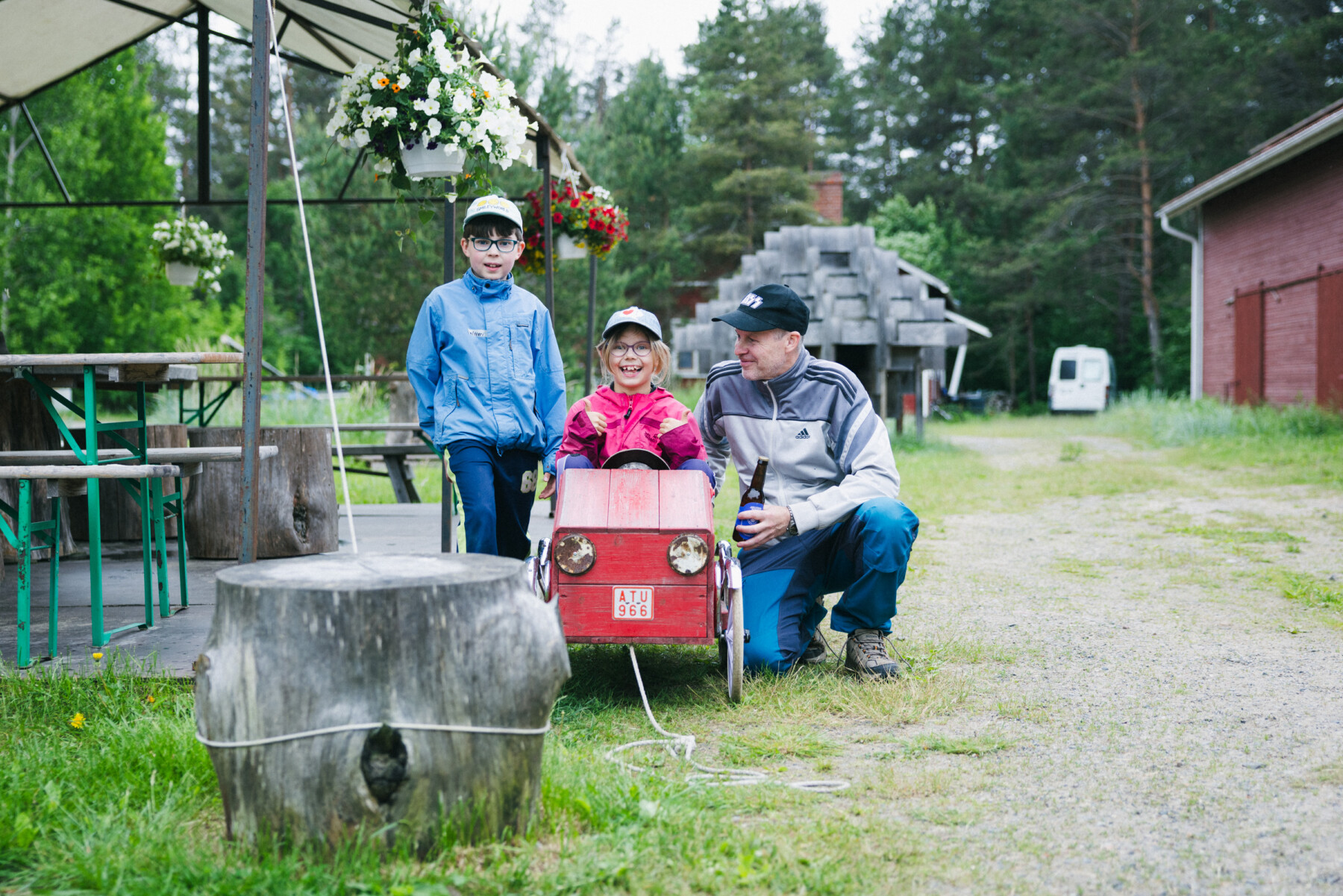 Two children and an adult pose beside a small red pedal car on a grassy path in Hailuoto, with trees, brick buildings and hanging flower baskets in the background.