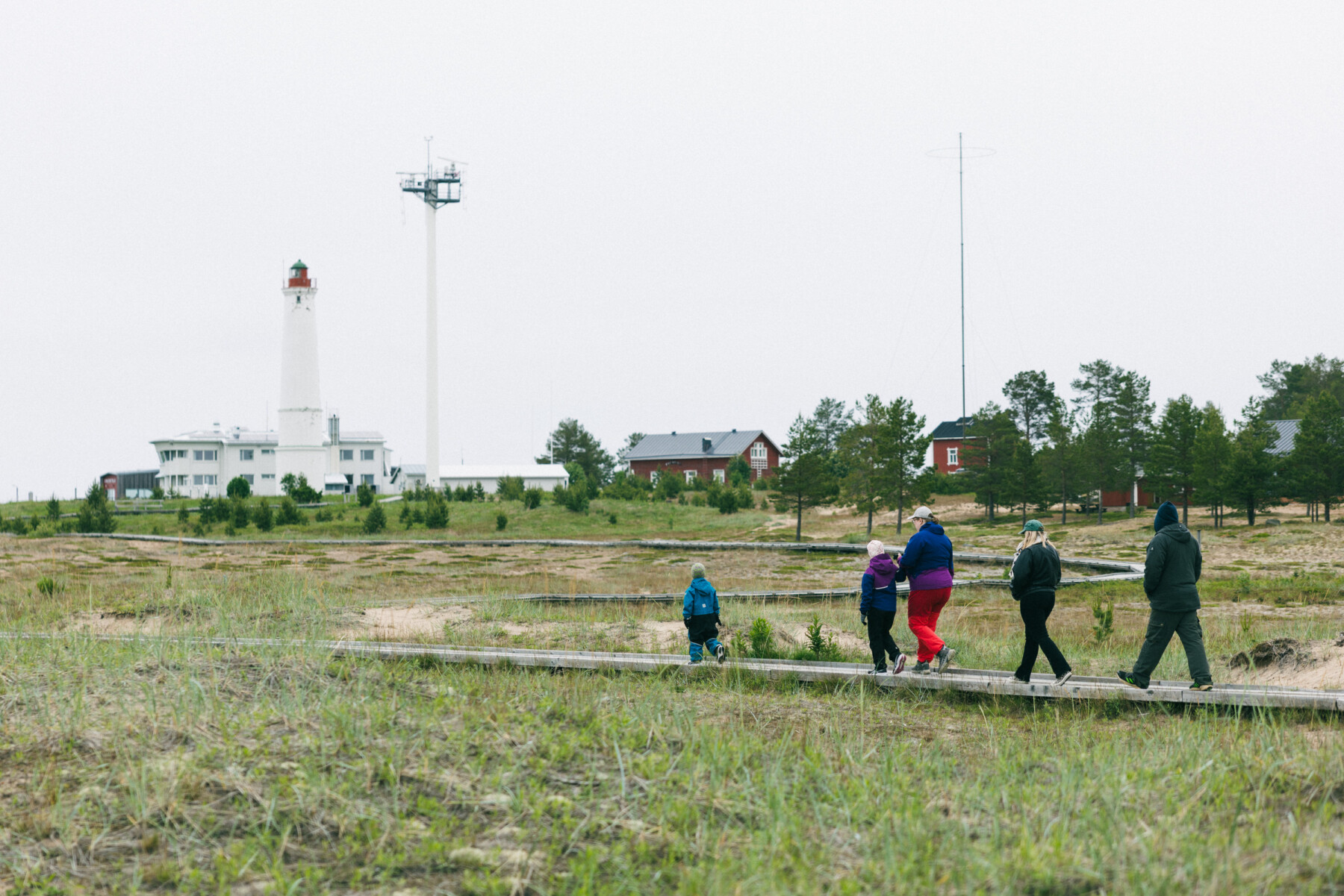 Visitors walk along a wooden boardwalk across coastal grassland, with the Marjaniemi Lighthouse and red wooden buildings visible in the background.
