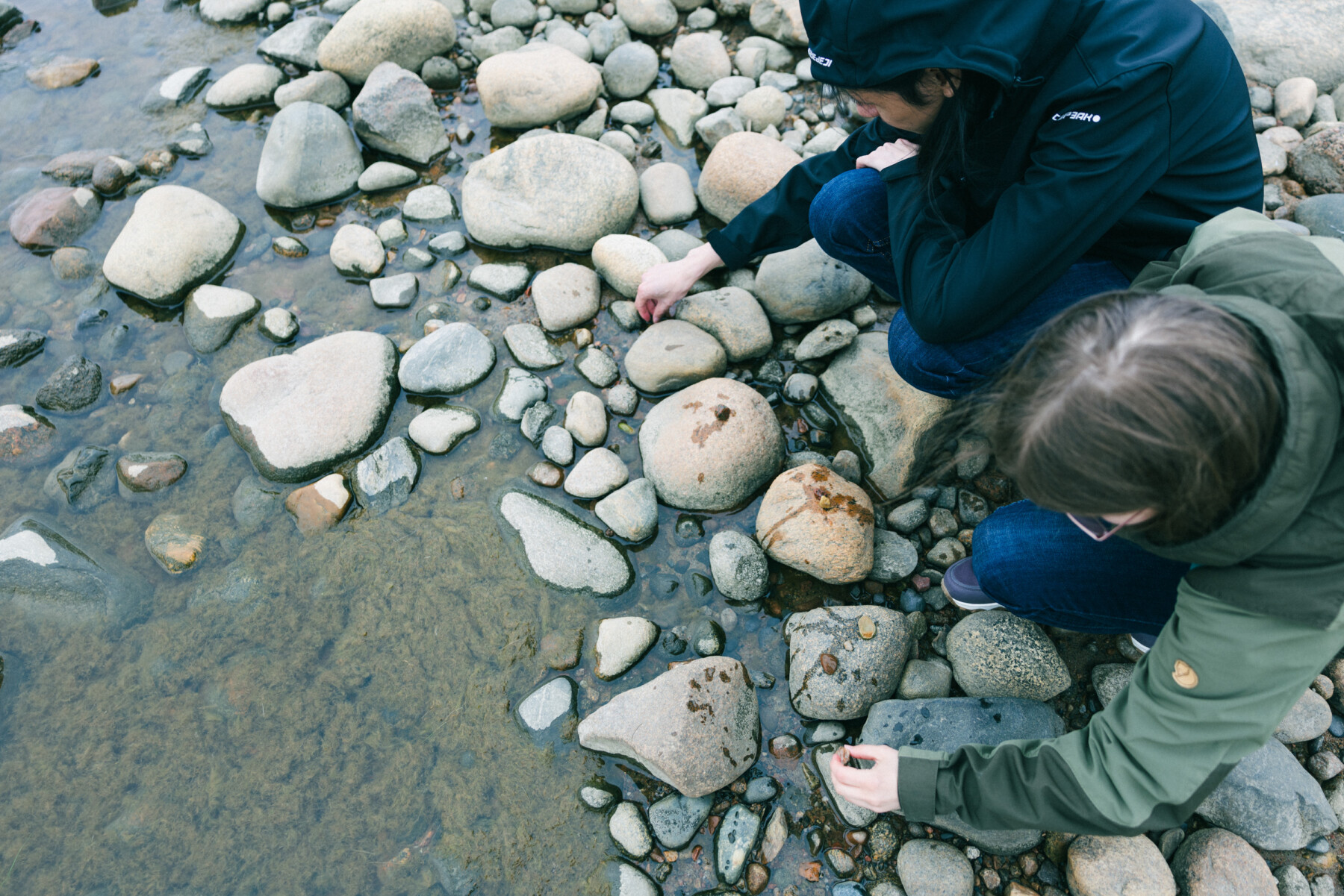 Two people kneel on a pebble-strewn shore, reaching towards small stones among larger rounded rocks at the water's edge.