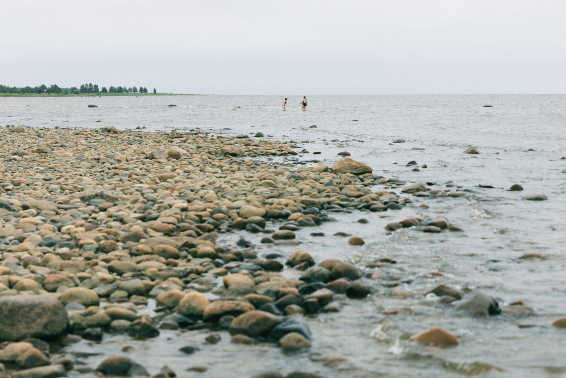 A rocky shoreline on Hailuoto stretches into shallow sea water, with two distant people standing in the water near the horizon under an overcast sky.