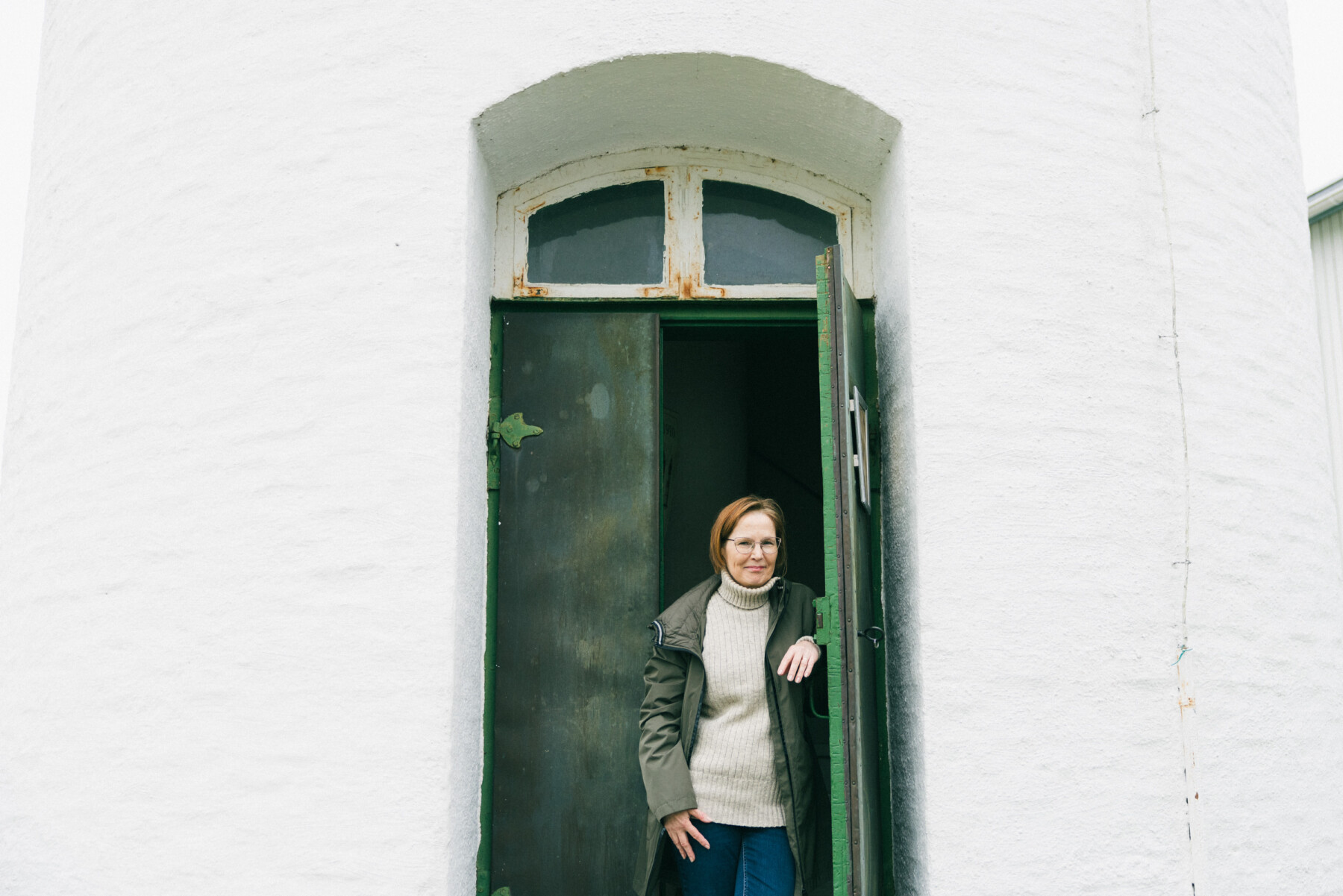 A woman wearing a light knitted jumper, dark jacket and glasses, leans against the entrance doorway of Marjaniemi Lighthouse, framed by a tall arched opening with dark green doors set into a white wall.