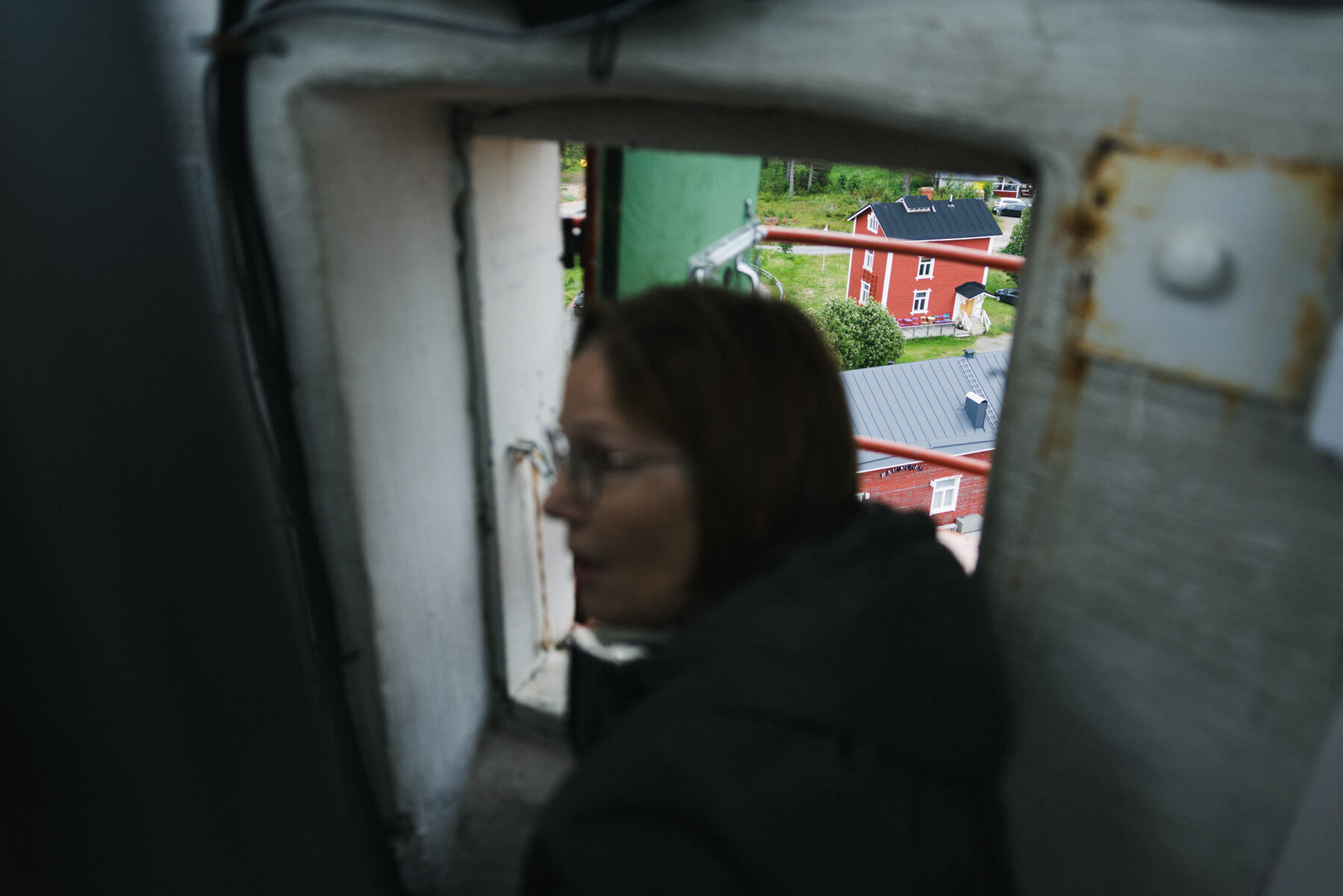 A woman climbs a narrow stairway inside Marjaniemi Lighthouse near a small hatch window, through which red buildings and part of the surrounding village are visible.