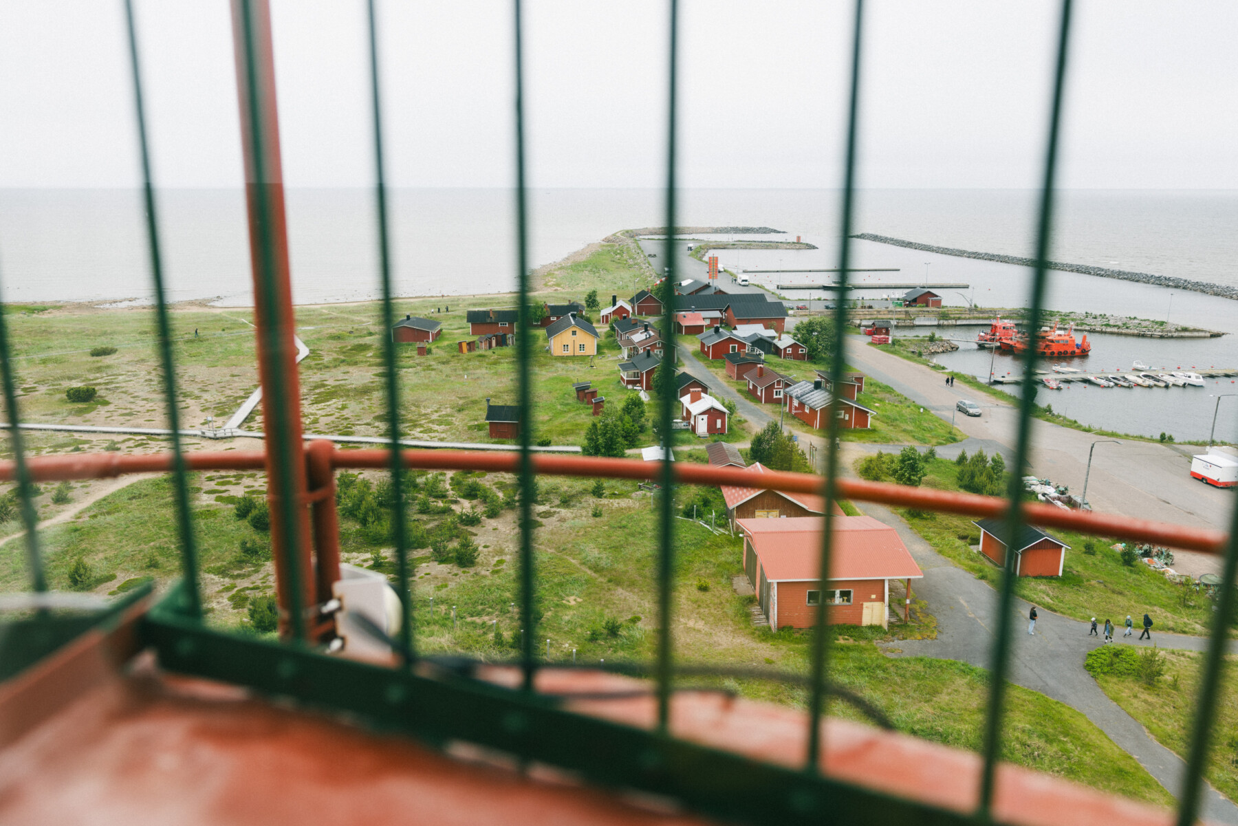Viewed through the safety railing of Marjaniemi Lighthouse, red buildings, a small harbour and breakwaters stretch along the shoreline beside the Gulf of Bothnia.
