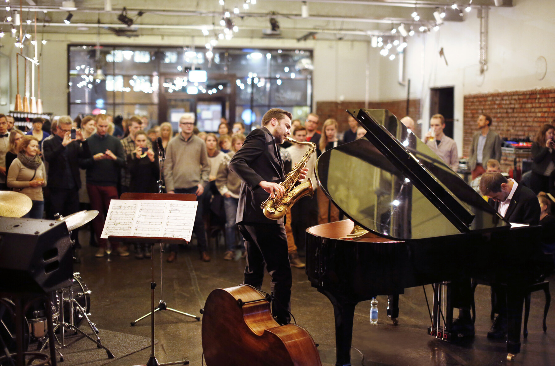 A man plays a tenor saxophone and another man plays a baby grand piano in front of a standing audience of several dozen people.