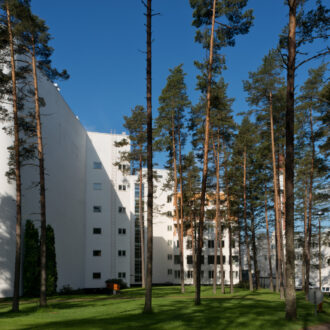 The large white Paimio Sanatorium stands behind a row of tall pine trees on a sunny summer day.