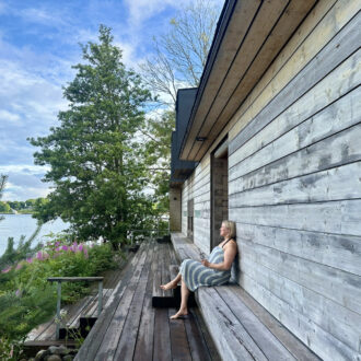 A woman sits on a wooden sauna bench wrapped in a towel. The summer landscape features nature and the sea.