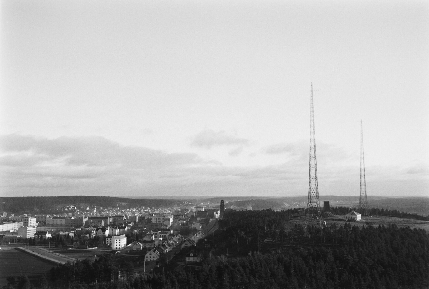 Two tall radio masts dominate the skyline on a hill above a city’s buildings, with countryside visible in the background.
