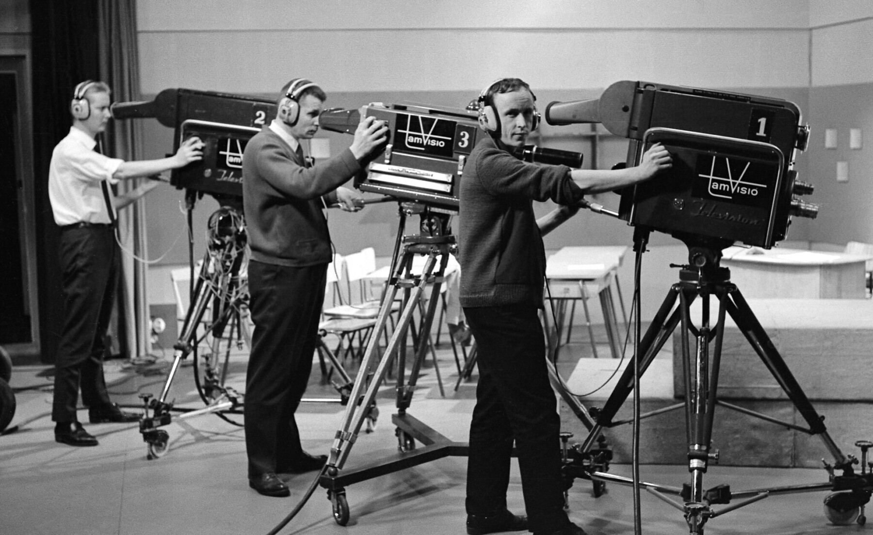 Camera operators wearing headphones pose behind three large studio television cameras labelled 1, 2 and 3, each mounted on a tripod.