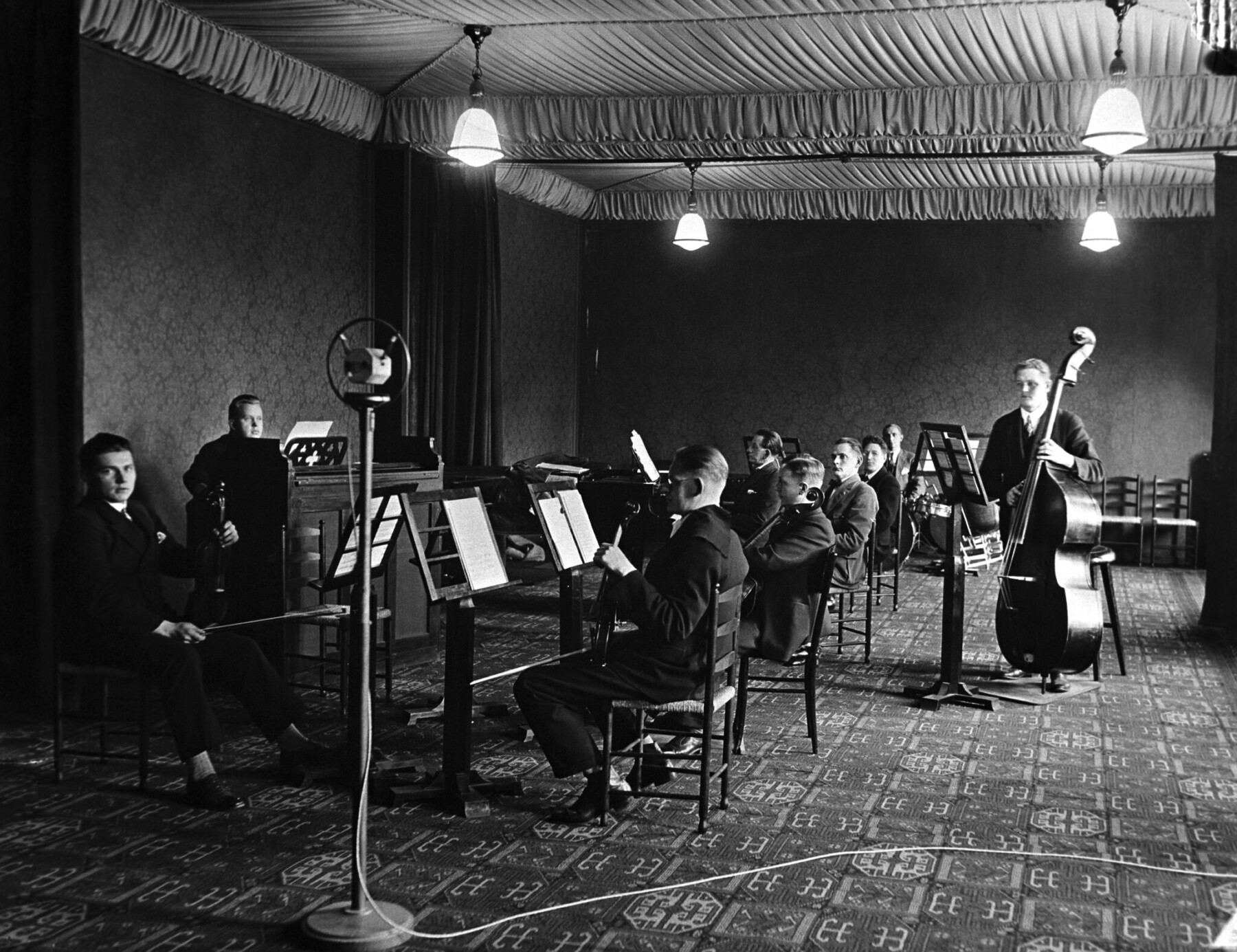 Finnish Radio Symphony Orchestra musicians perform in a room with hanging lamps, seated behind music stands that hold sheets of paper.