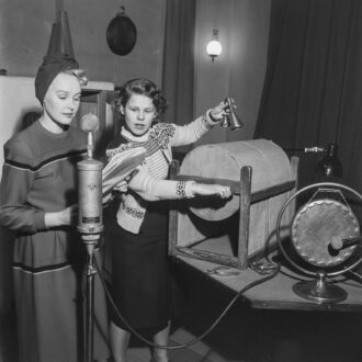 Actor Toini Vartiainen, left, reads from a script beside sound engineer Marja Aaltonen, who operates a large wooden drum and holds two small bells, during a 1949 Yle radio broadcast.