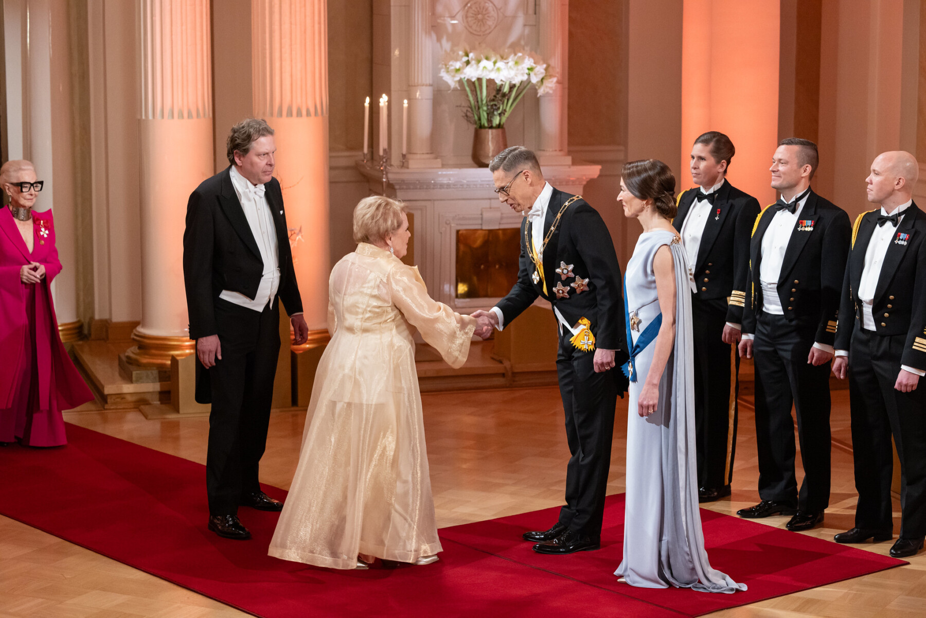 Katri Helena, wearing a long cream-coloured gown, shakes hands with President Alexander Stubb on a red carpet at the Presidential Palace in Helsinki.