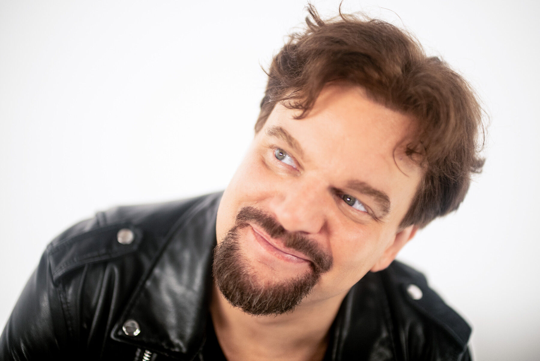 Ismo Leikola poses in a black leather jacket and a goatee, glancing upwards with a slight smile against a white background.