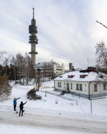 A winter view of Helsinki shows a cylindrical concrete tower that rises above the surrounding buildings, while two people walk their dogs through the snow.