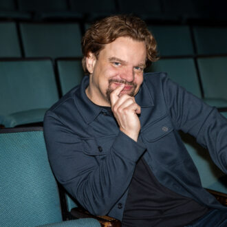 Finnish comedian Ismo Leikola sits among empty theatre seats, resting his chin on one hand as he looks towards the camera with a smile.