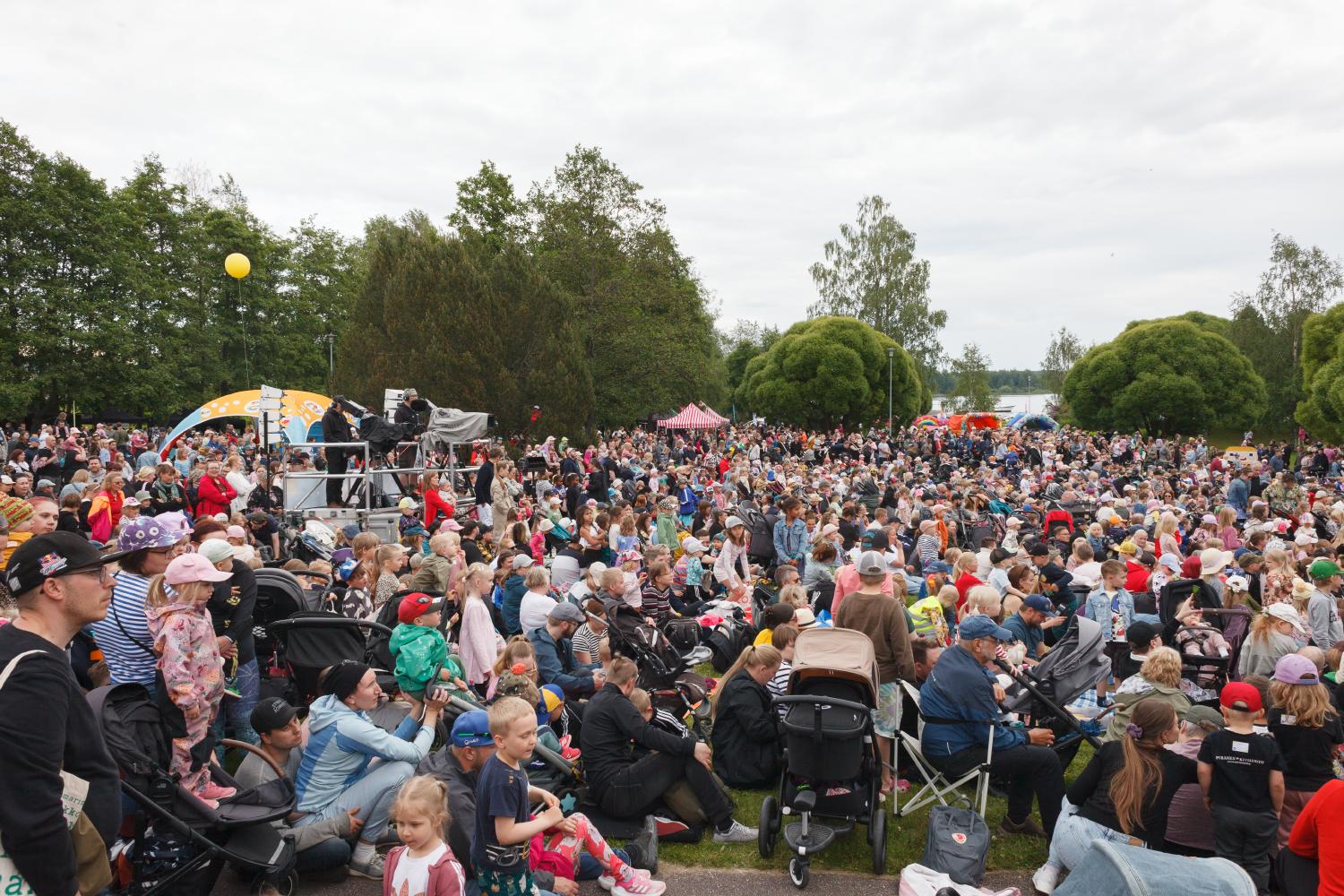 Families and children fill a grassy park in Laukaa for Yle’s Pikku Kakkonen concert, with rows of people and folding chairs surrounded by trees.