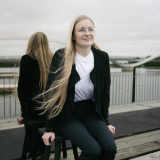 Armi Ruusuvirta sits on a stool outside Olosauna, with the Oulu River and a bridge reflected in a mirror behind her.
