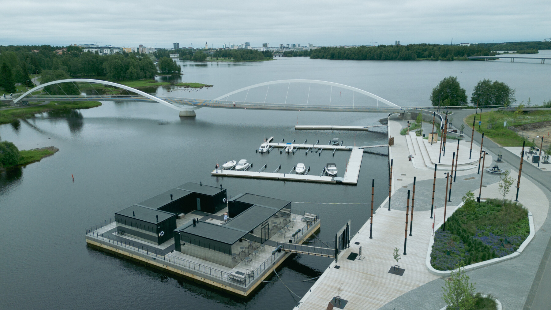 Olosauna sits on floating pontoons on the Oulu River, with its open courtyard beside a marina, pedestrian paths and an arched bridge.
