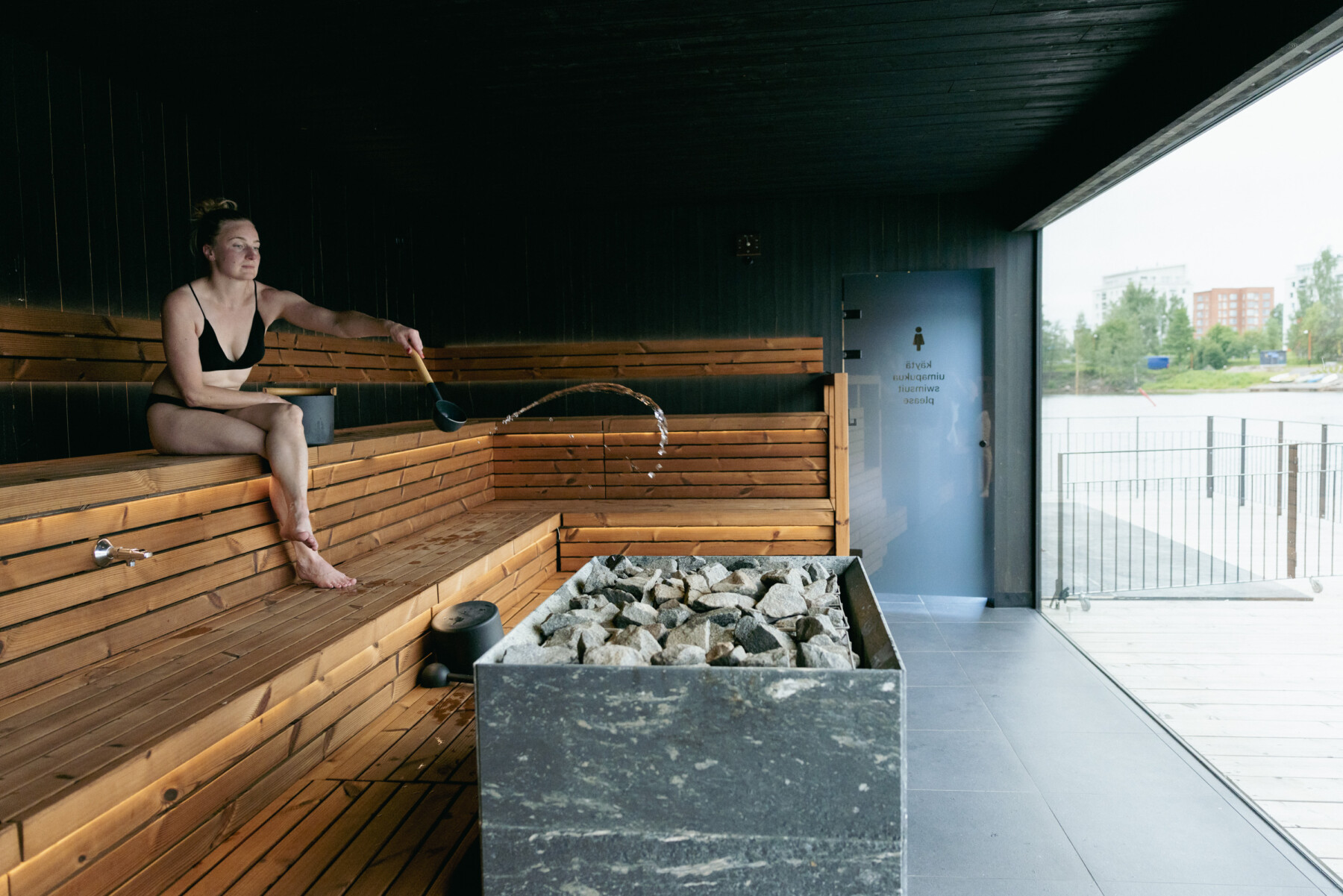 A woman sits on the tiered wooden benches of a sauna in Oulu, tossing water from a ladle onto the sauna stove.
