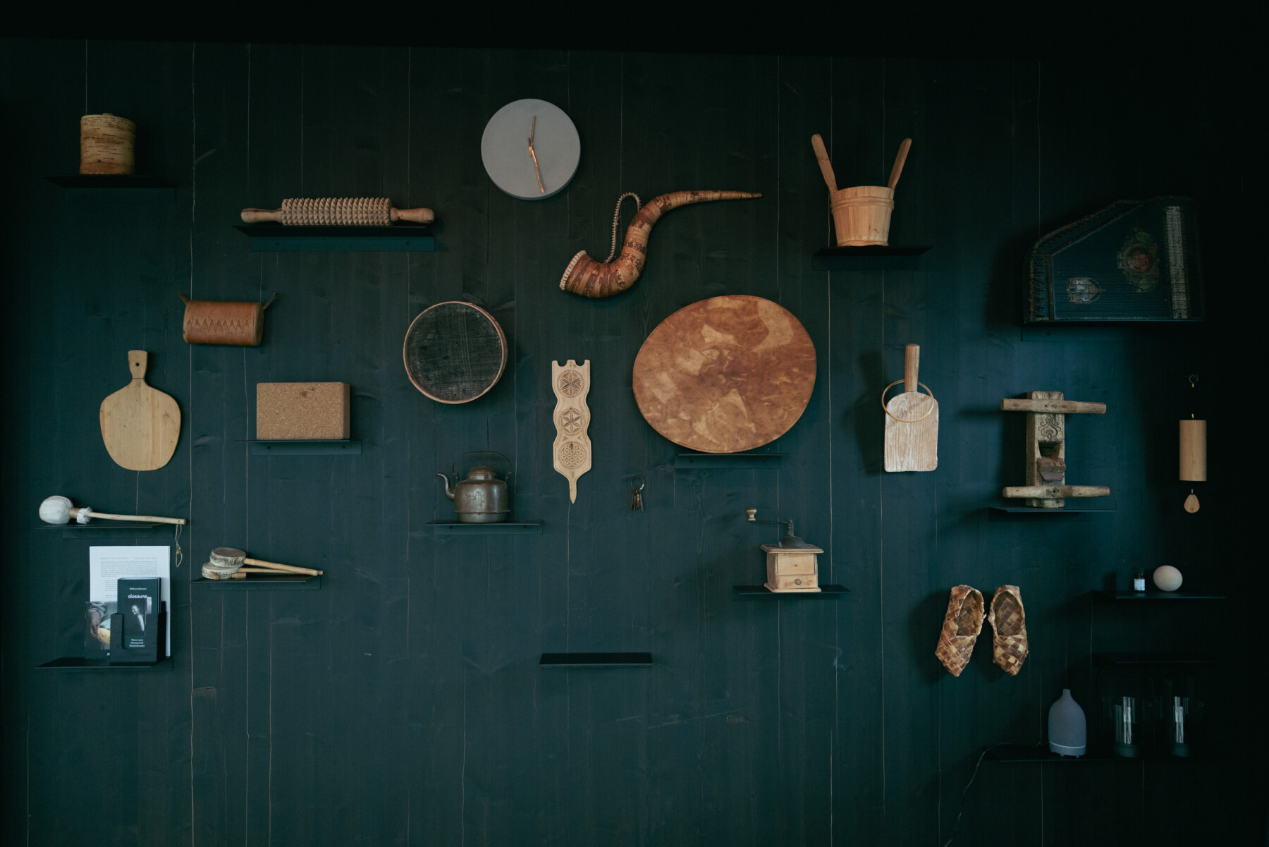 A collection of traditional Finnish household objects and handcrafted wooden items is displayed on shelves on a dark green wooden wall.