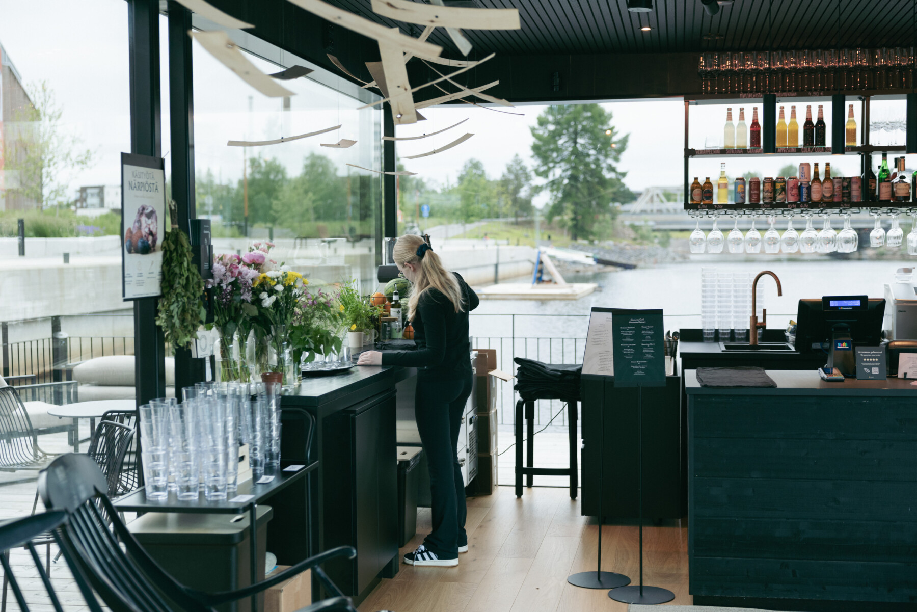 A woman stands at the Olosauna café counter, surrounded by flowers, glassware, bottles and drinks, with the waterfront visible through the windows.
