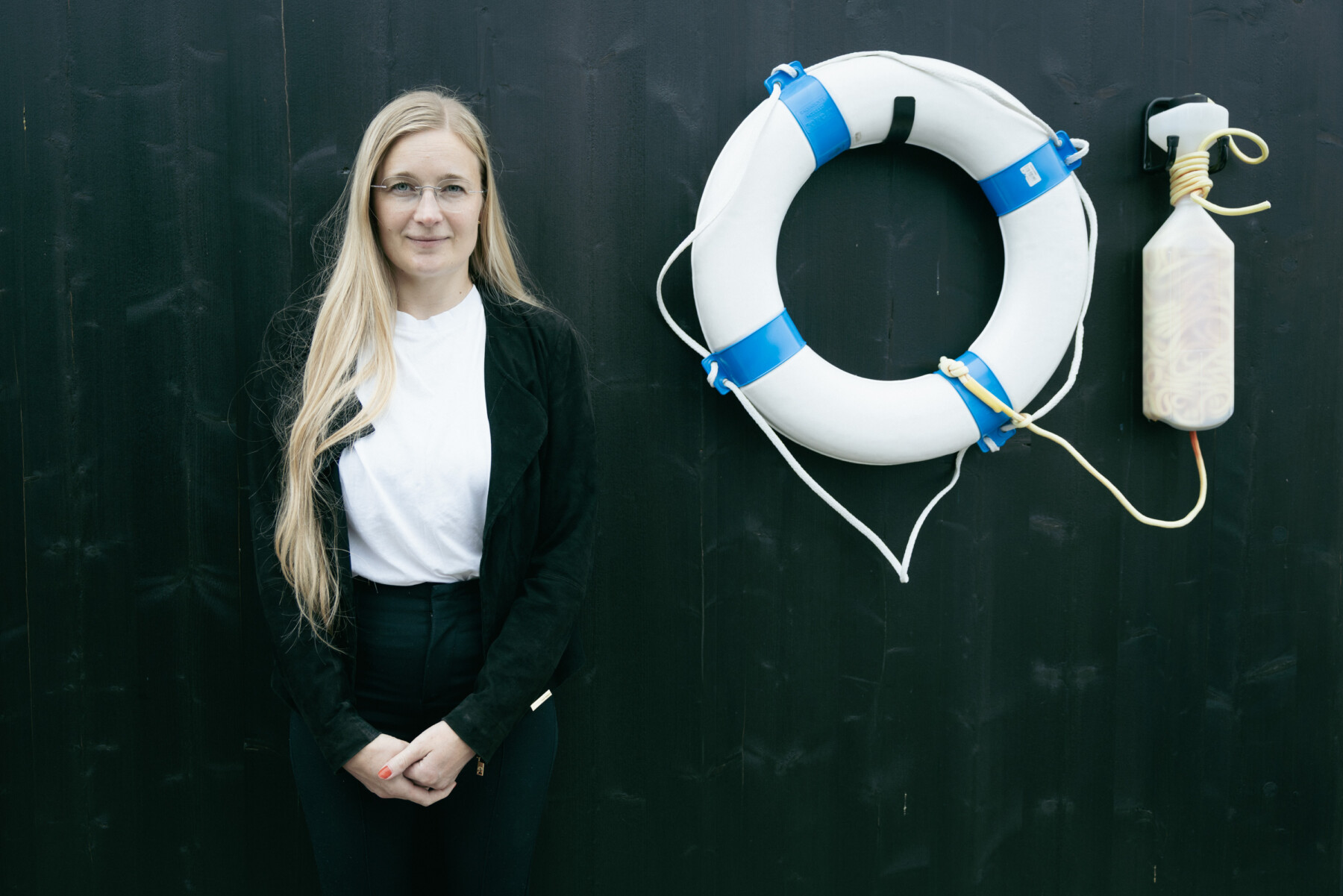 Armi Ruusuvirta stands against a dark wall beside a white life ring with blue bands and a white rope-tied float.