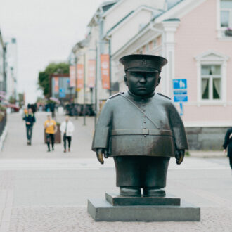 The Bobby at the Market Place statue by Kaarlo Mikkonen depicts in bronze a stocky police officer in uniform and cap standing on a low stone plinth in Oulu Market Square, with people and historic buildings visible behind it.
