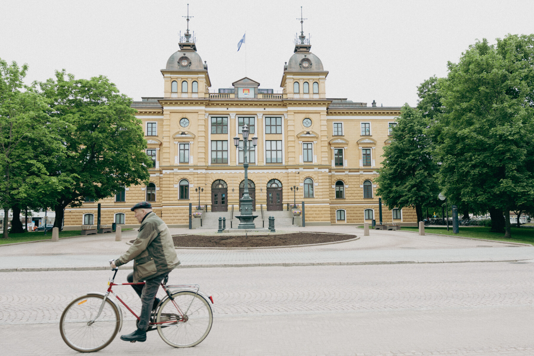 A cyclist rides across a cobbled square in front of Oulu City Hall, a yellow historic building framed by large leafy trees.