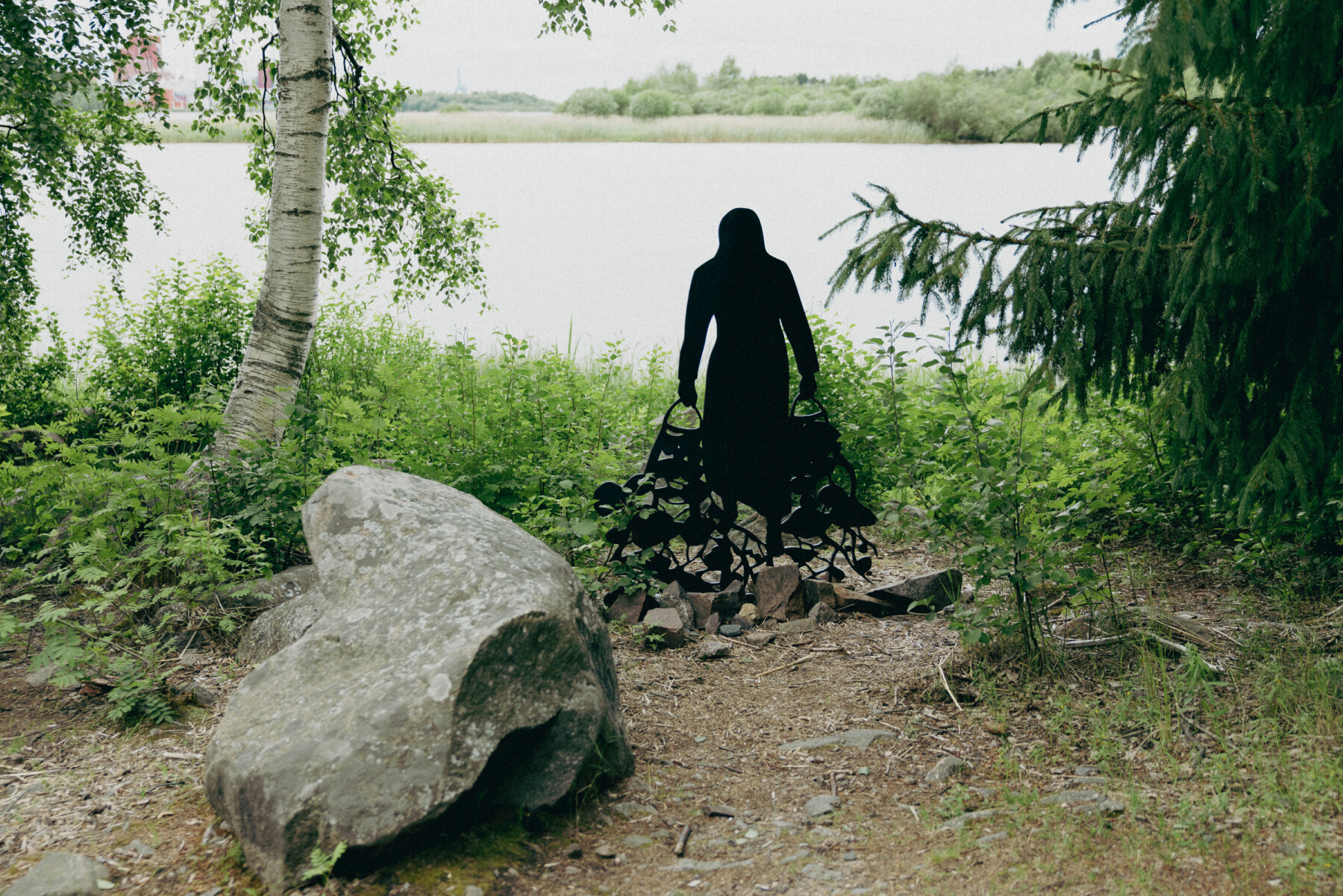A black silhouette sculpture from Jane Remm’s Hunch, Care and Thirst of Life stands at the edge of a lakeshore, surrounded by trees, shrubs and rocks.