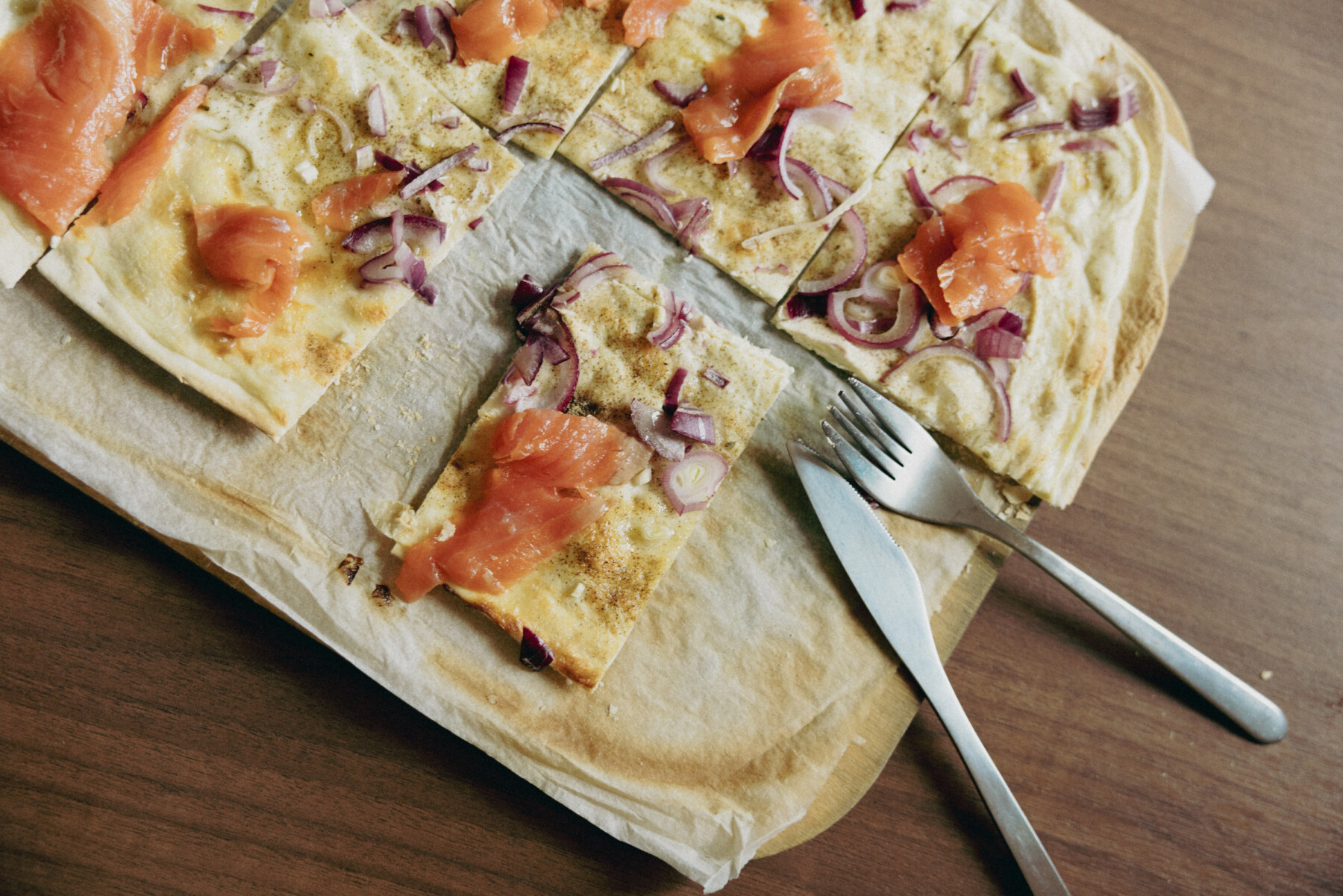 A thin-crust Flammkuchen topped with smoked salmon and red onion is sliced into rectangles and served on baking paper with cutlery alongside.