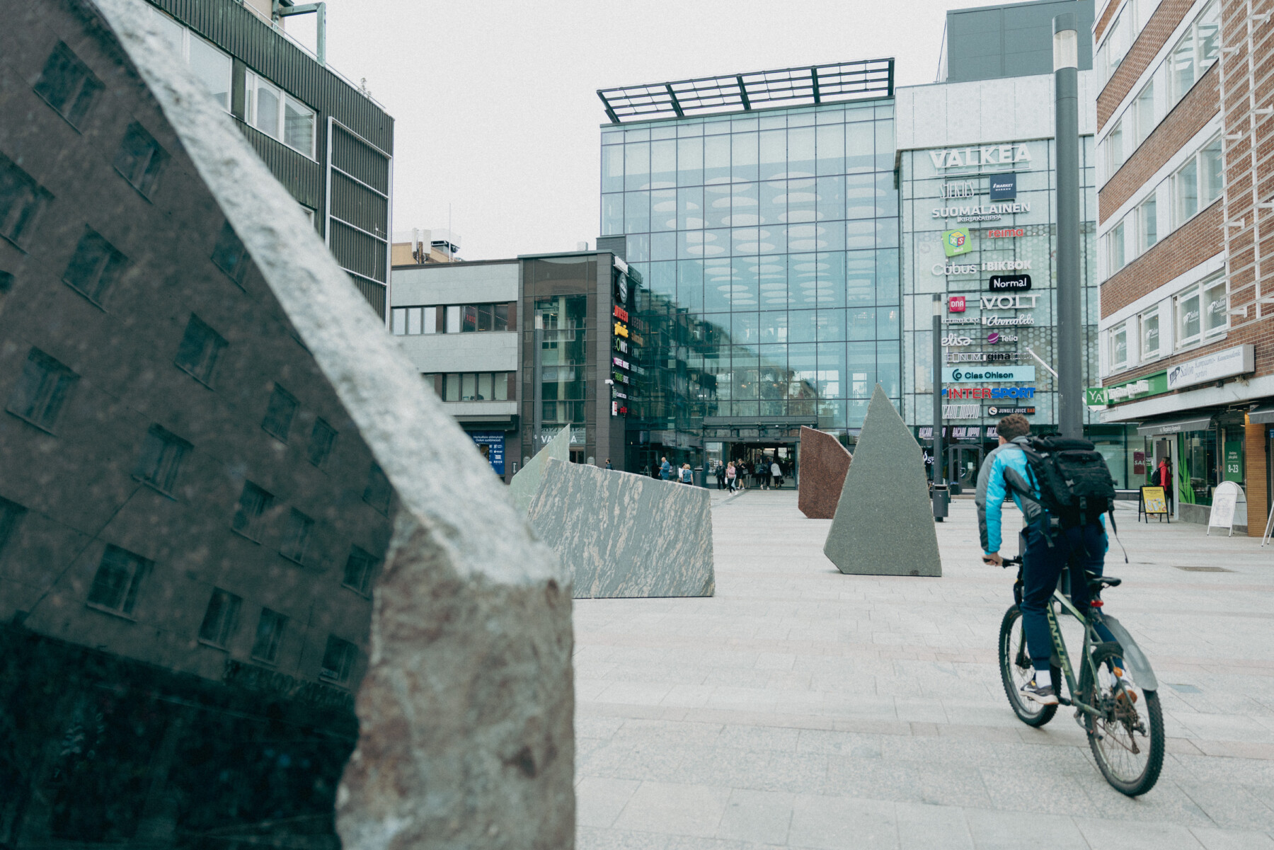A cyclist rides past the angular stone sculptures of Rana Begum’s No. 1574 Stone in a paved square surrounded by modern buildings in central Oulu.