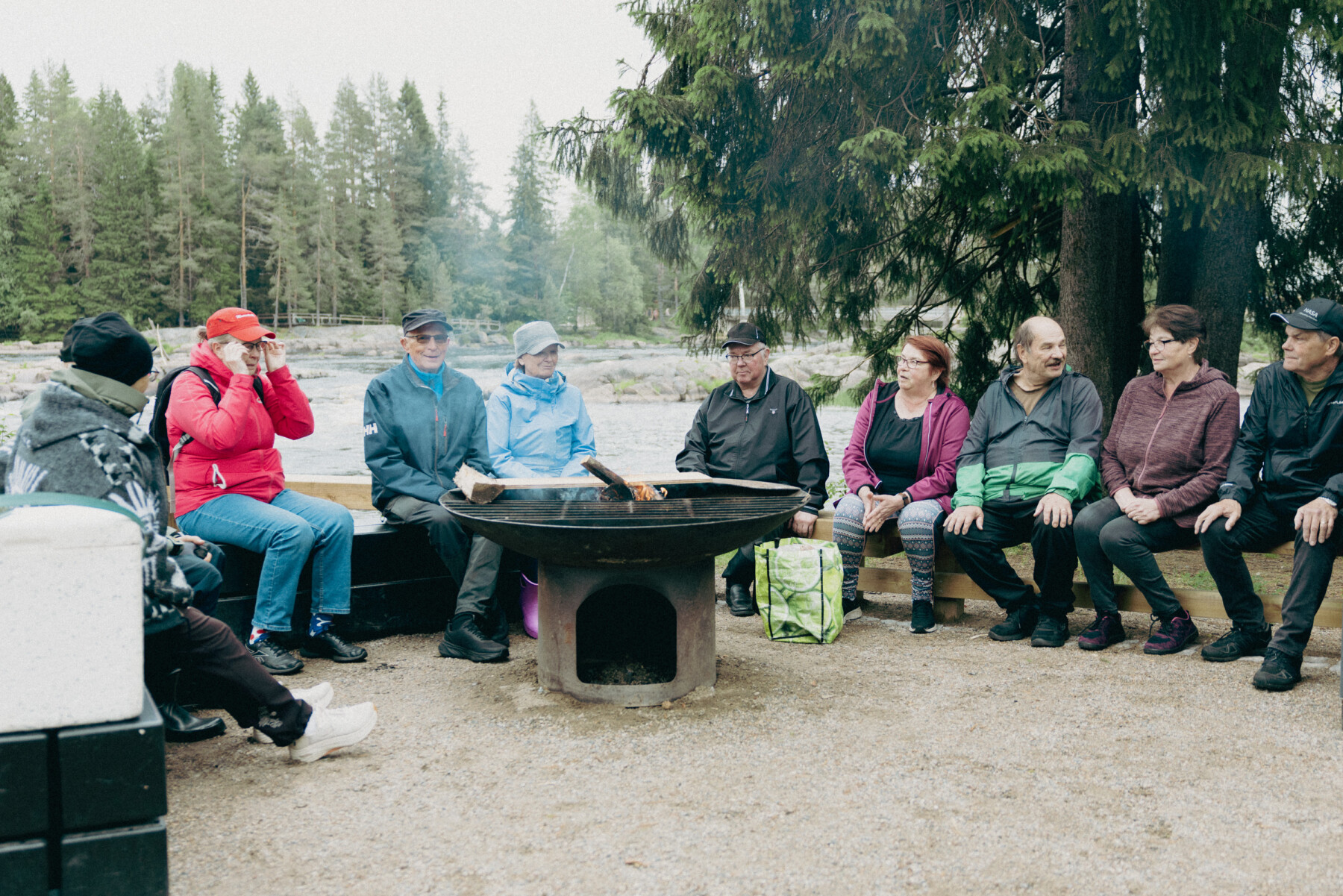 A group of people in jackets and hats sits around a circular fire pit beneath tall pine trees, with a river flowing behind them.
