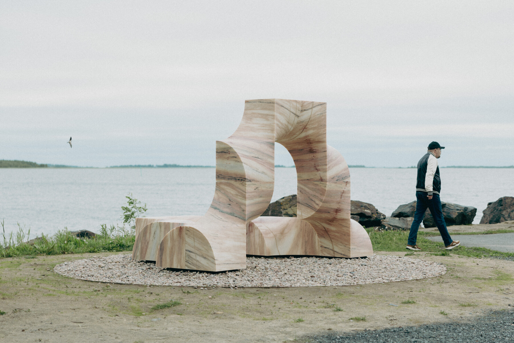 Superflex’s Super Kello features stacked curved stone forms on a circular gravel base beside the waterfront at Kiviniemi fishing harbour in Haukipudas, with a person walking past.