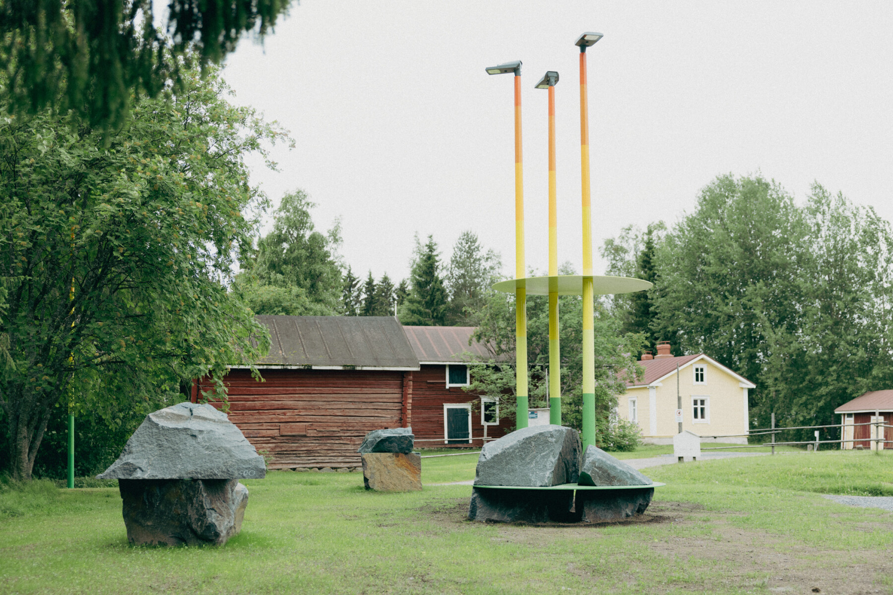 Three slender poles with green-to-yellow-to-red gradients rise above large granite boulders in Gabriel Kuri’s Risk Assessing Risk Assessment.