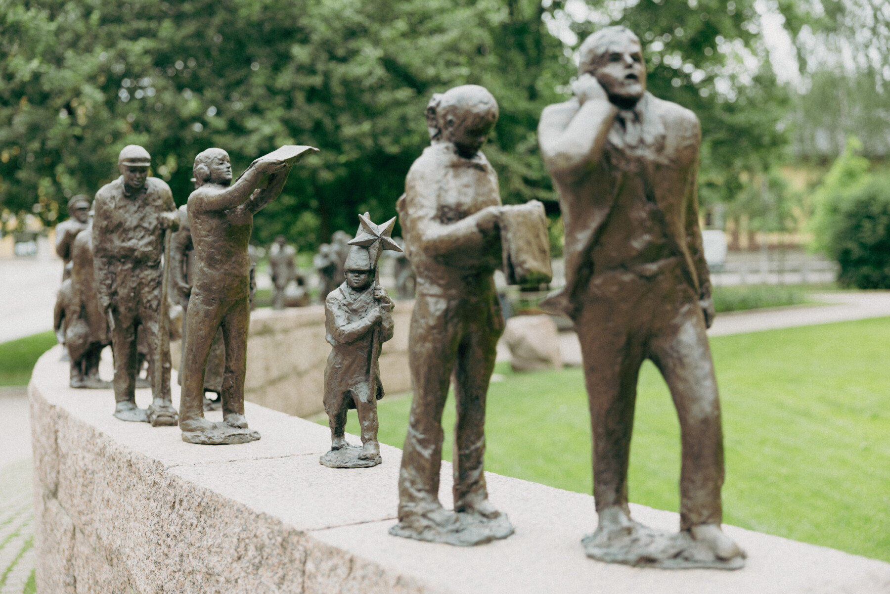 The Passage of Time sculpture by Sanna Koivisto features a line of bronze figures of different heights standing along a low stone wall in a leafy park.