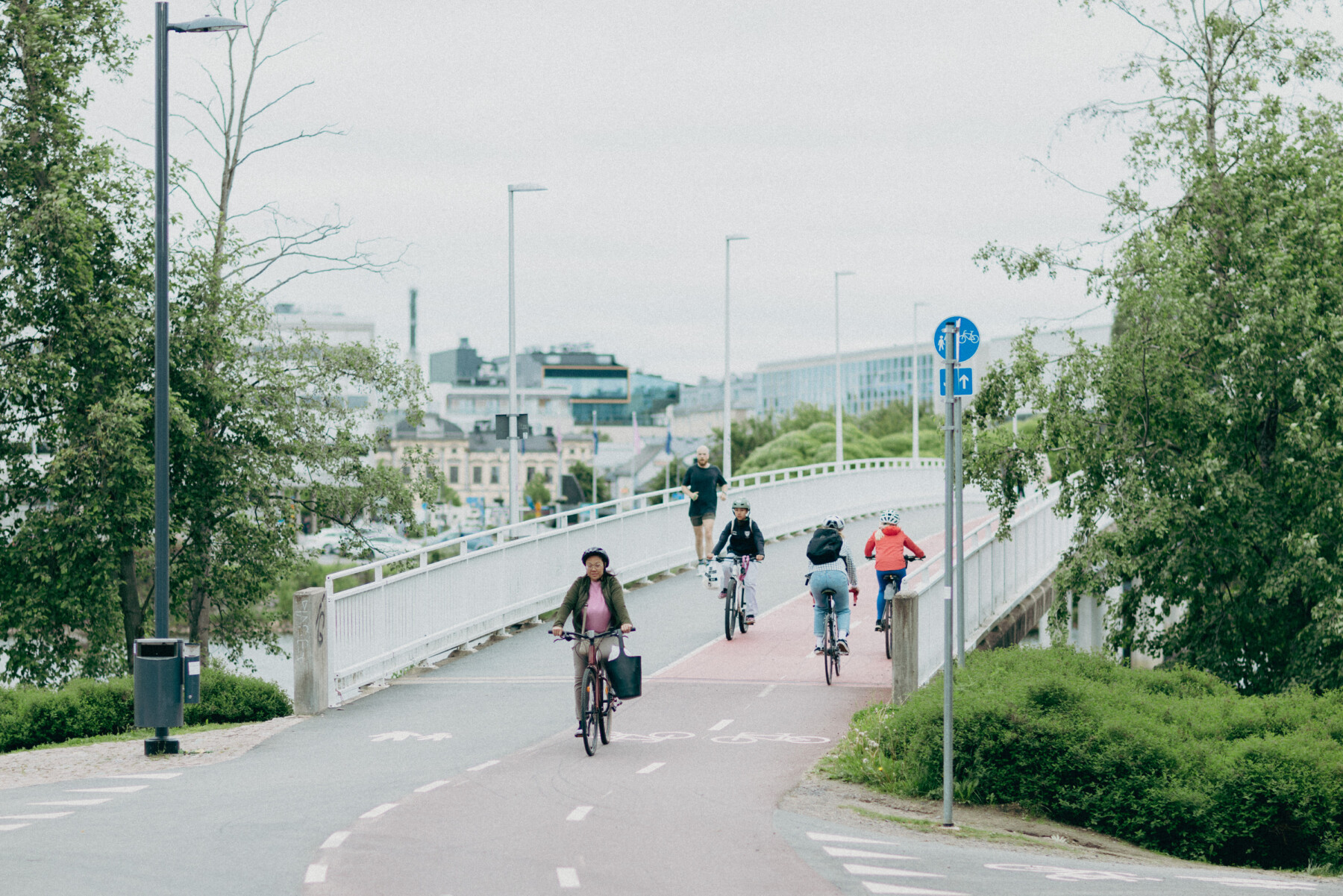Cyclists and a pedestrian cross a bridge on one of Oulu’s cycle routes, with trees and city buildings visible in the background.
