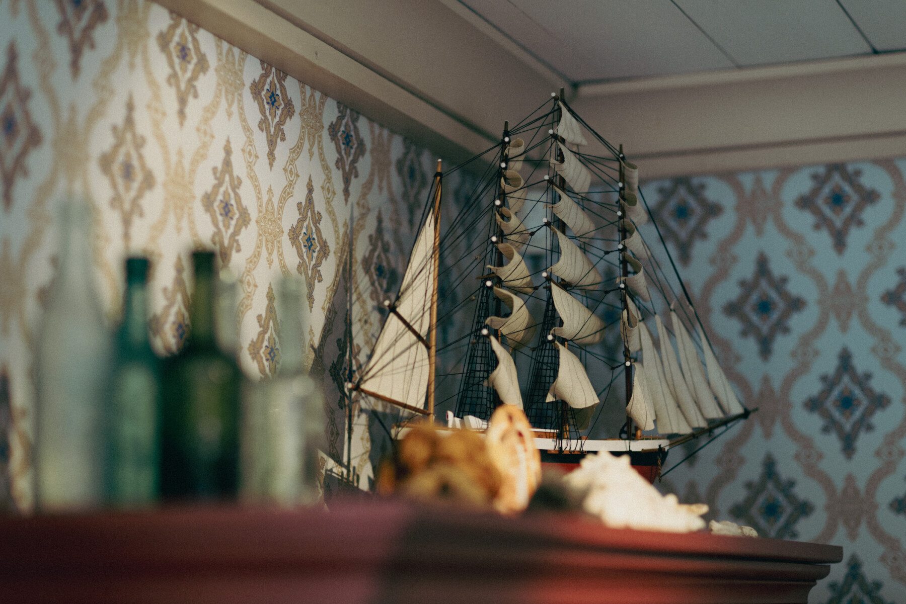 A detailed model sailing ship sits on a wooden cabinet inside the Sailor’s Home Museum, with patterned wallpaper, glass bottles and seashells in the foreground.