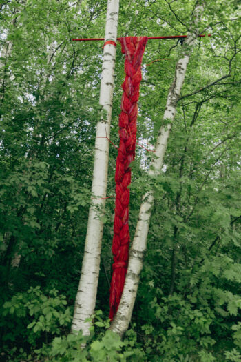 Helena Kaikkonen’s Plait features a long red braided textile suspended between white birch trees in a dense green forest.