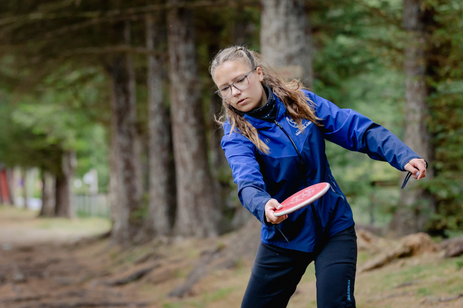 A female athlete in a blue jacket, Silva Saarinen, starts a throw with a red disc on a wooded forest path.