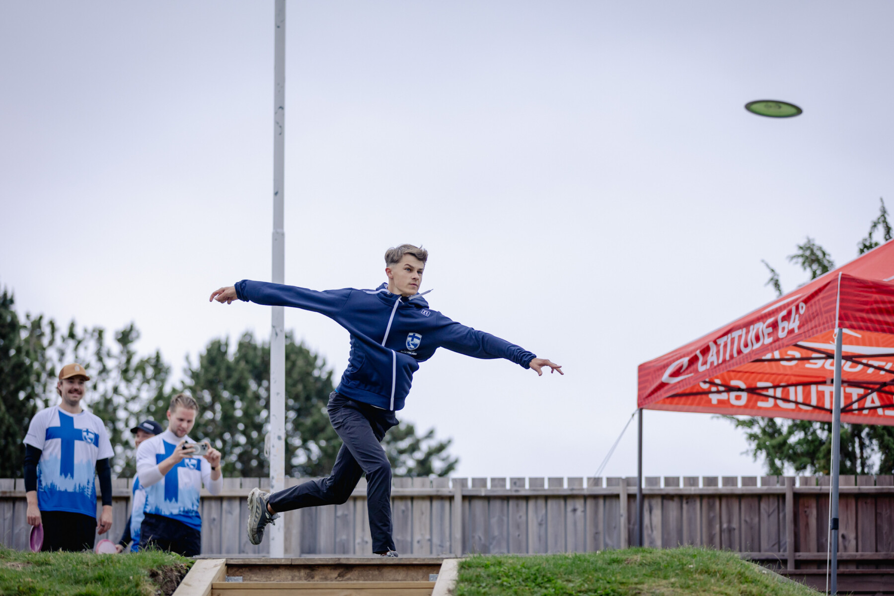A male athlete in a tracksuit, Niklas Anttila, extends his arms after releasing a disc, watched by two spectators in matching Finland jerseys nearby.