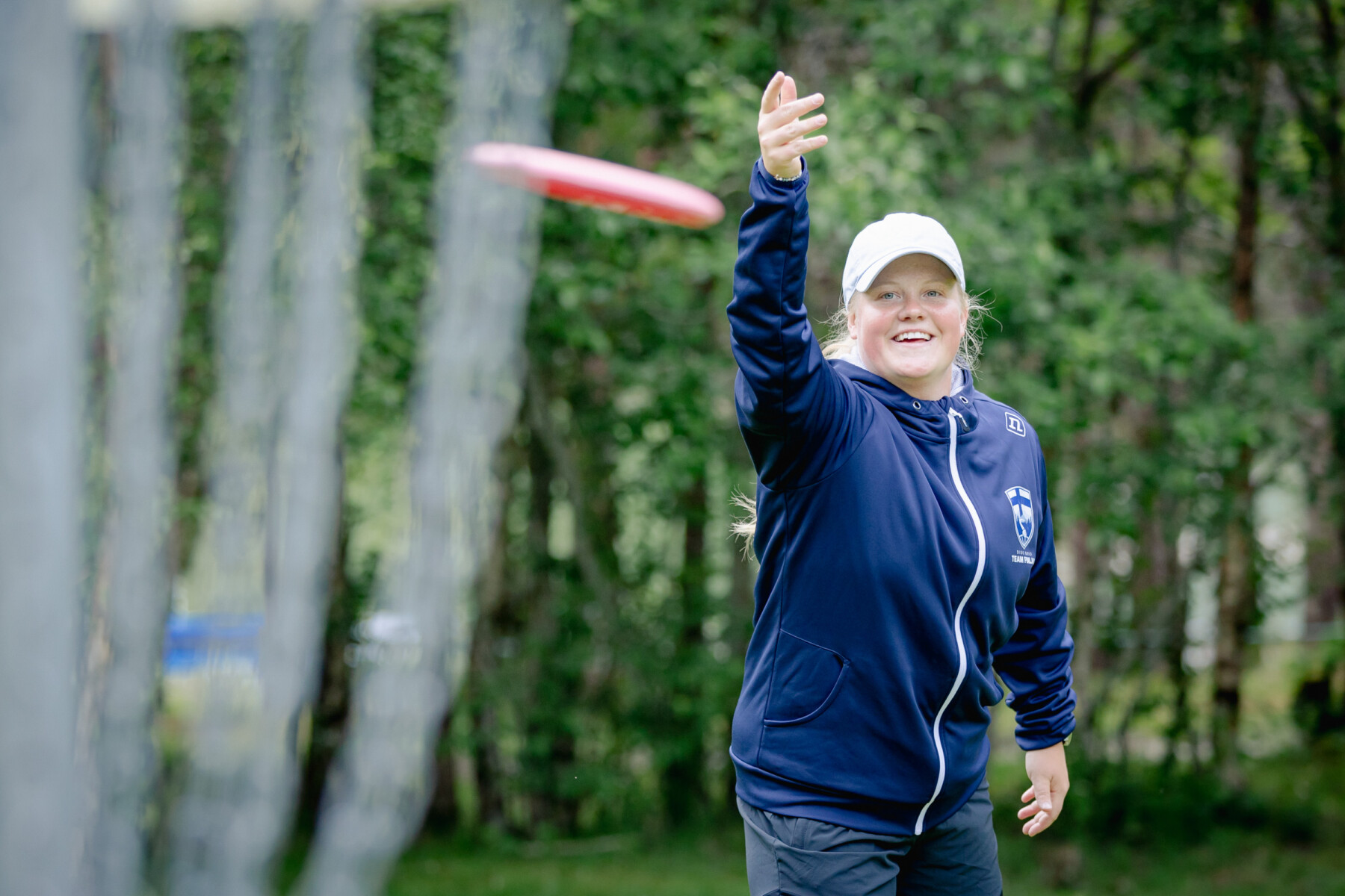 A smiling woman in a white cap and blue Team Finland jacket, Eveliina Salonen, has a raised arm after releasing a red disc towards the camera.