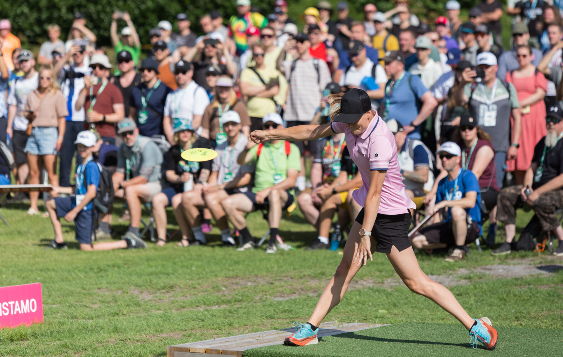 A female athlete in a pink polo shirt and black cap throws a yellow disc while spectators look on from the grass behind her.
