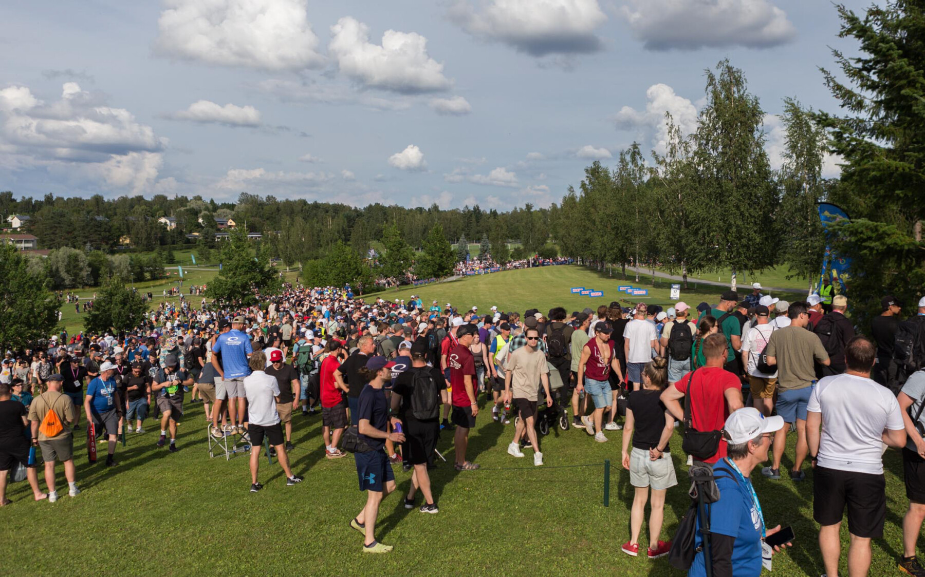 A crowd of spectators in summer clothing gathers on a sloping grass fairway lined with trees during an outdoor sporting event.