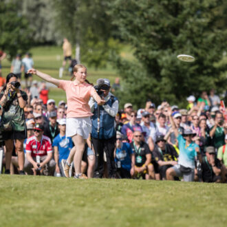 An athlete in a peach polo shirt and braids, Iida Lehtomäki, watches her disc fly past a large crowd of spectators.
